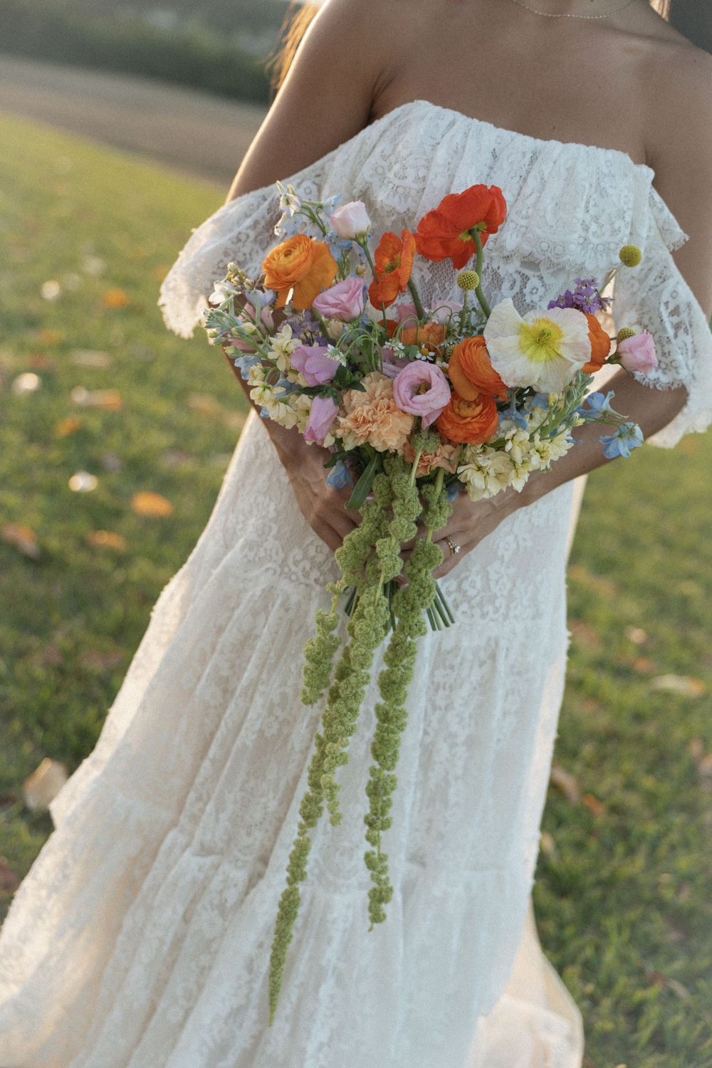 Bride in white lace off-shoulder gown holding colorful wildflower bouquet with cascading greenery outdoors.