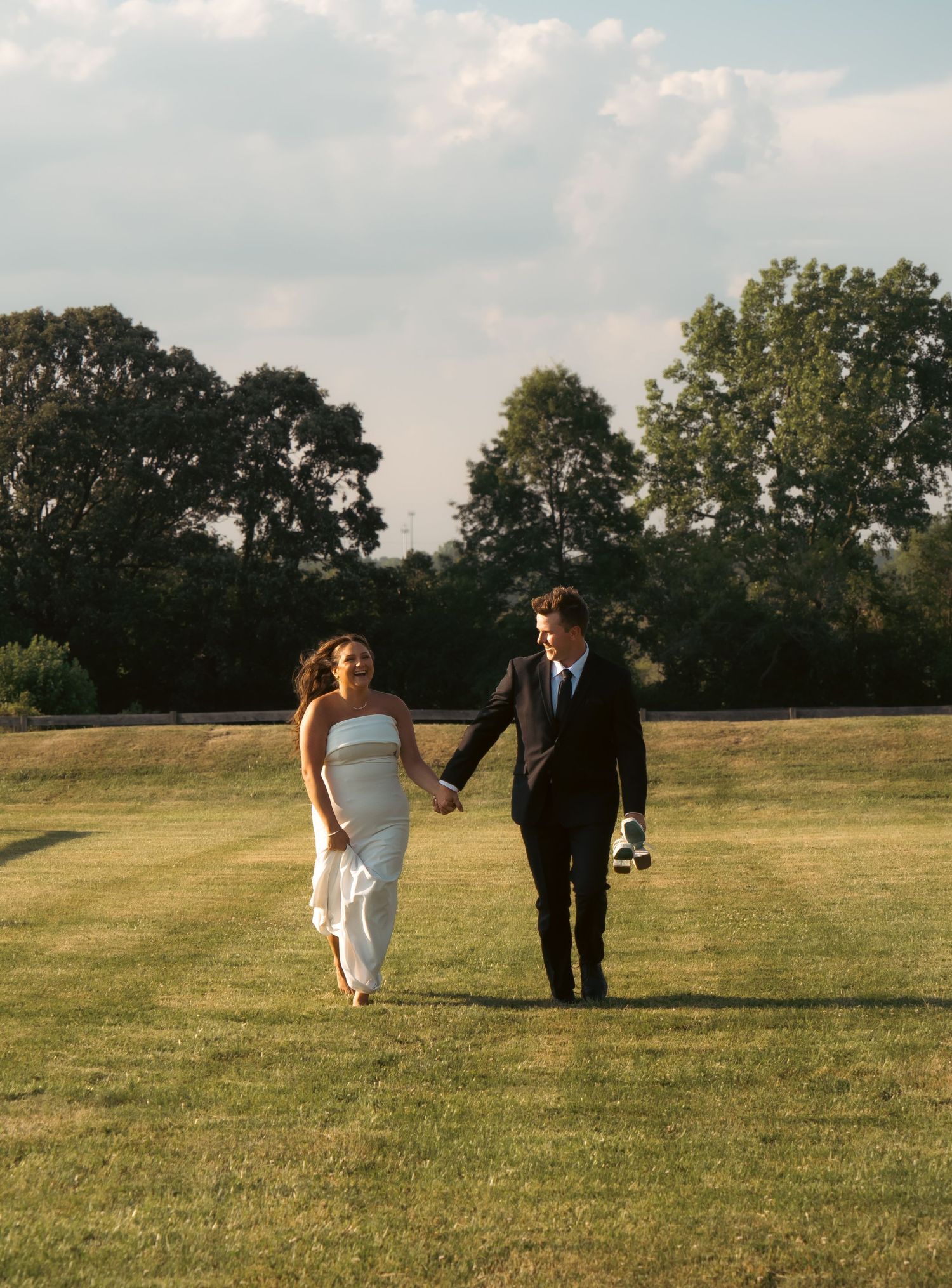 A couple walks hand in hand across a scenic grassy field on their wedding day at sunset.