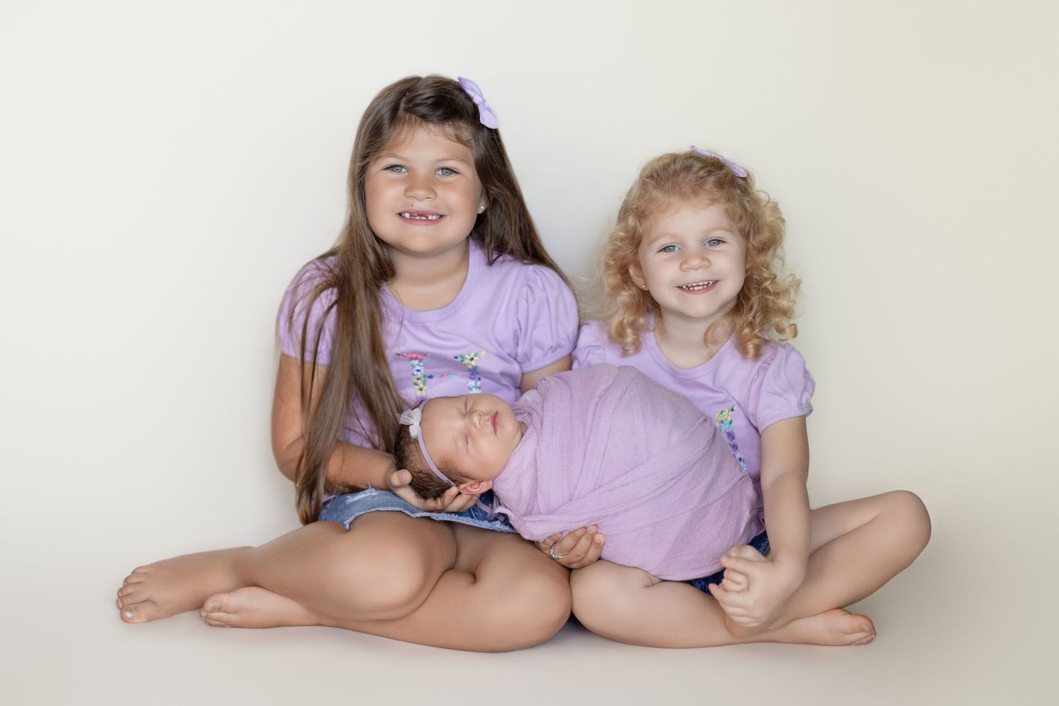 Three siblings in purple outfits sitting together on a beige background during a studio photo session.