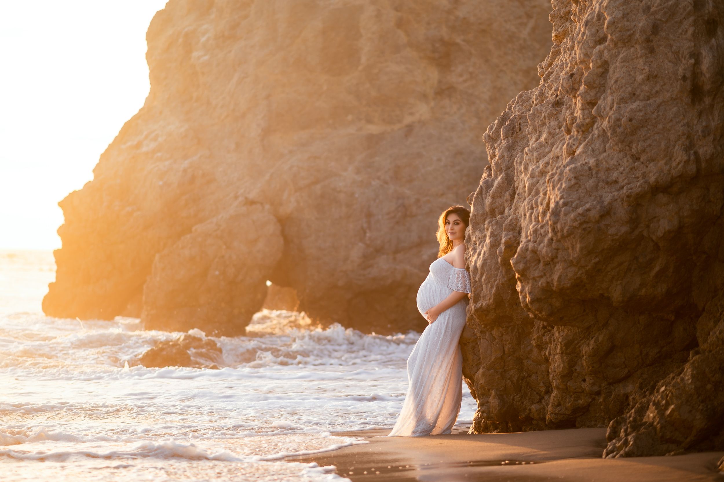 Pregnant woman in flowing white gown standing by ocean cliffs bathed in warm golden sunset light.