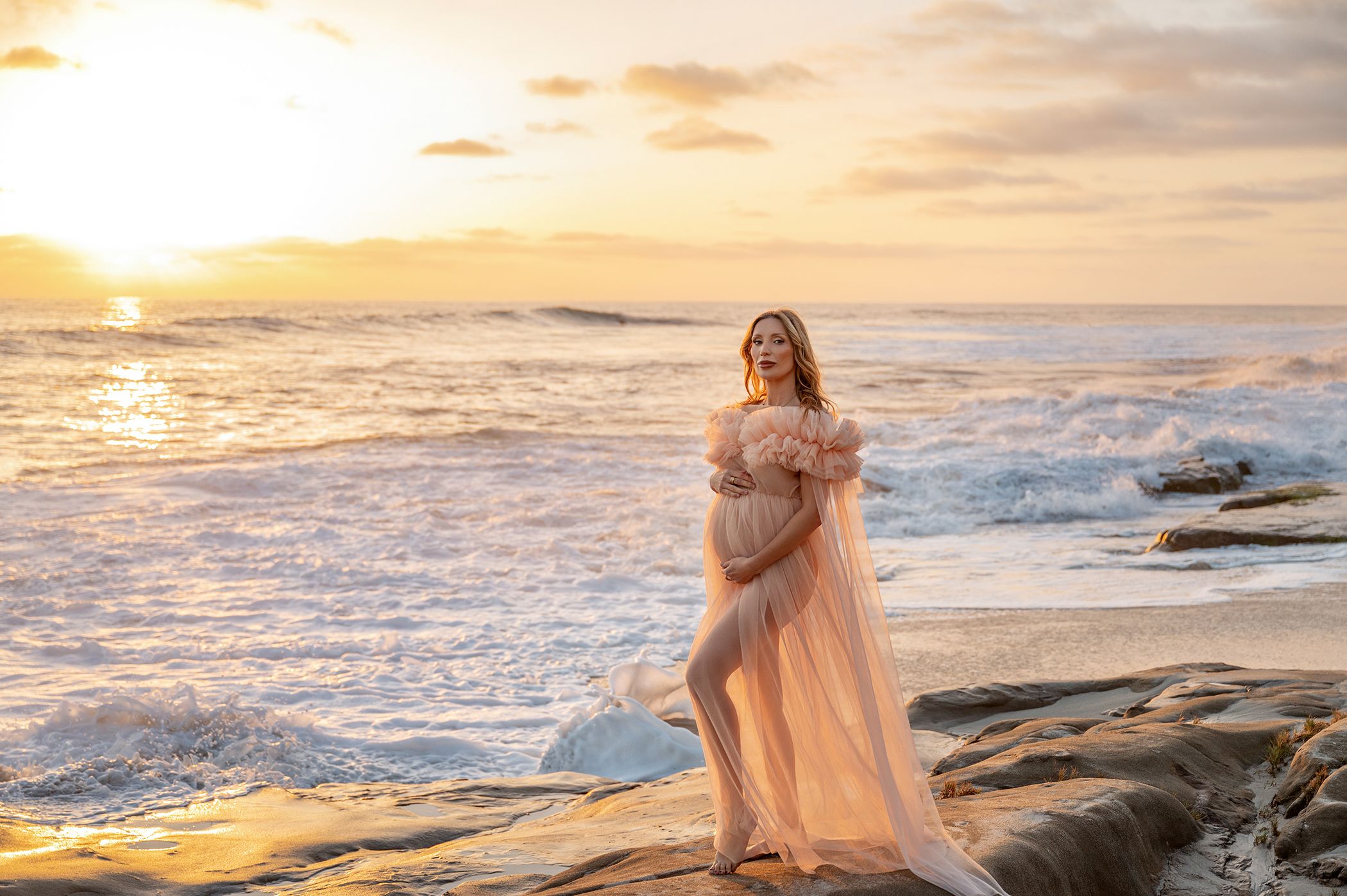 Pregnant woman in sheer peach gown stands on rocks at golden sunset beach.
