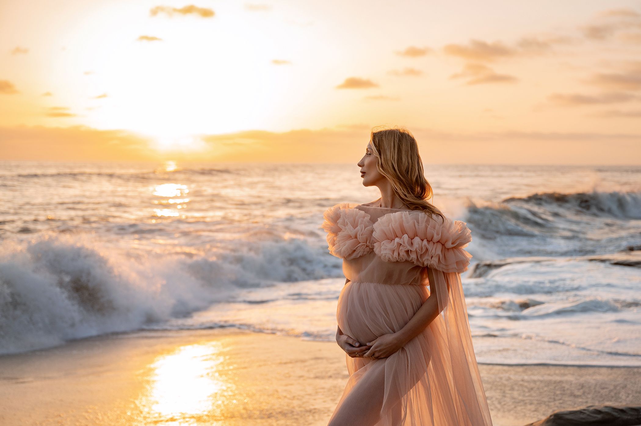 A pregnant woman in a blush tulle gown cradles her bump at sunset on a rocky beach.