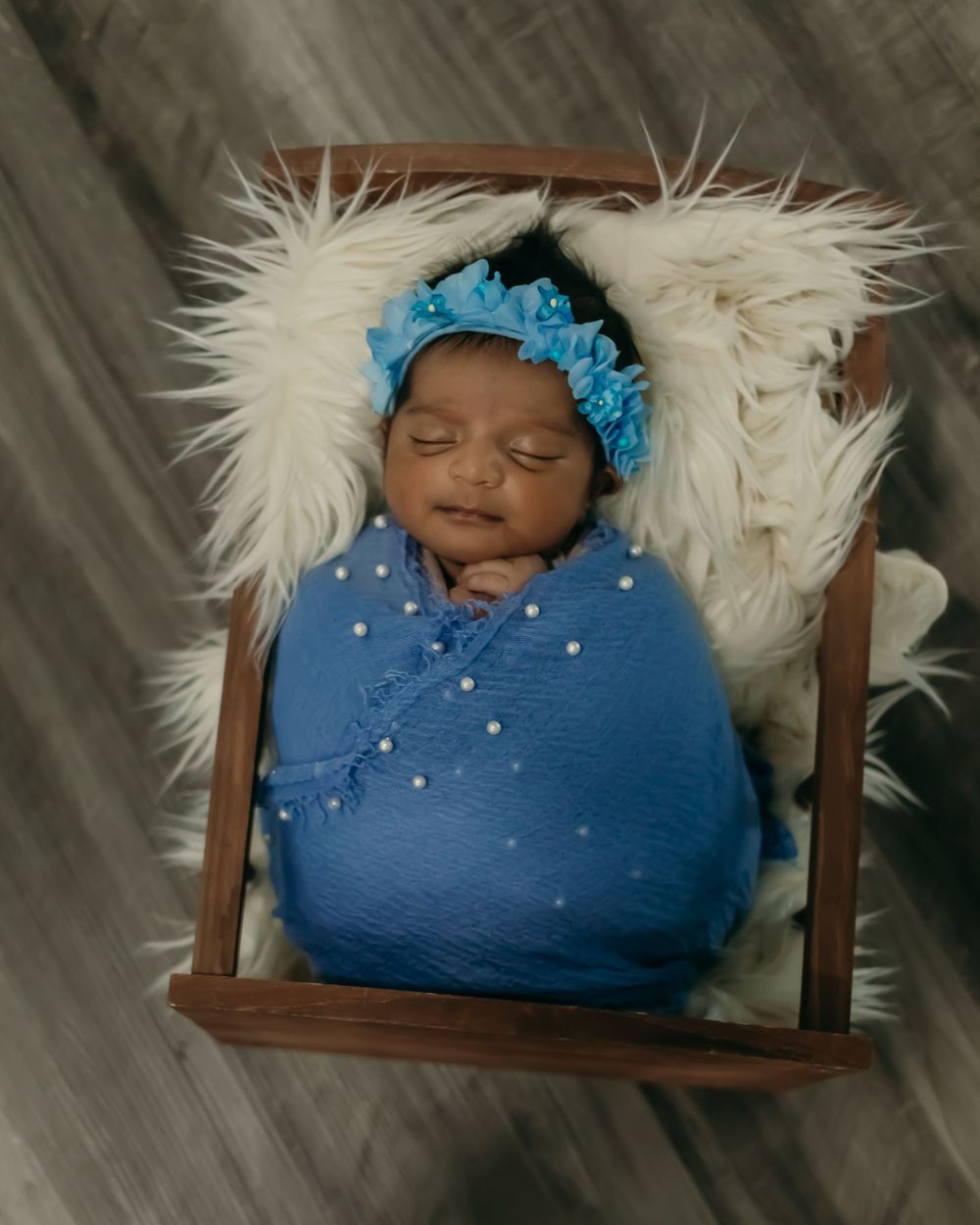 Sleeping newborn wrapped in blue fabric with a turquoise floral crown rests on white fur in a wooden prop bed.