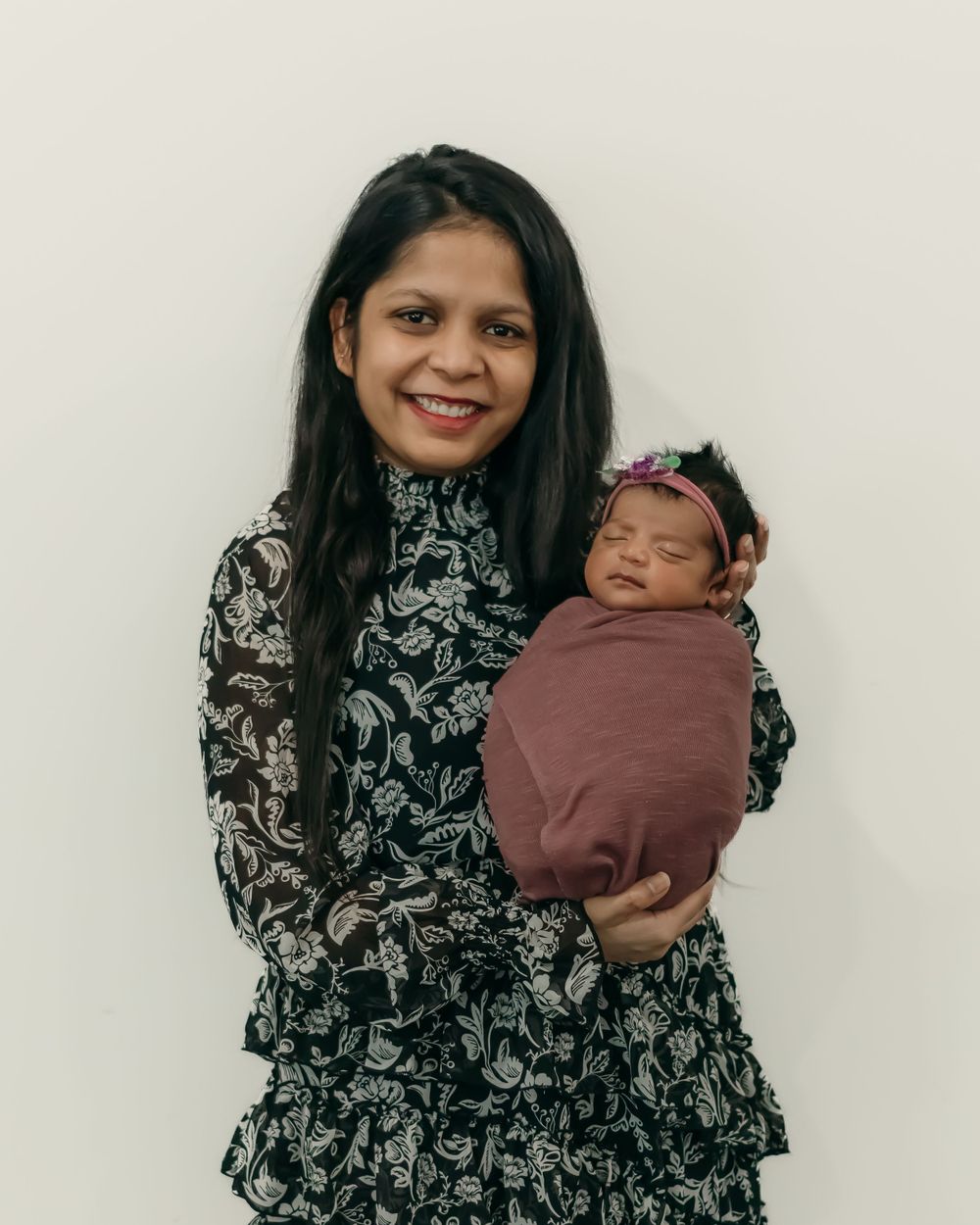 Woman in floral dress holds swaddled infant against white wall.