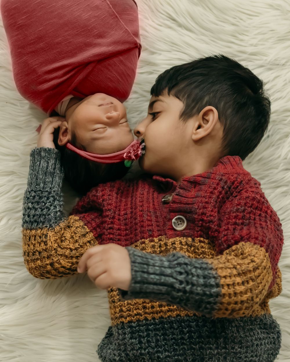 Two siblings in winter sweaters share tender moment on white fur blanket.