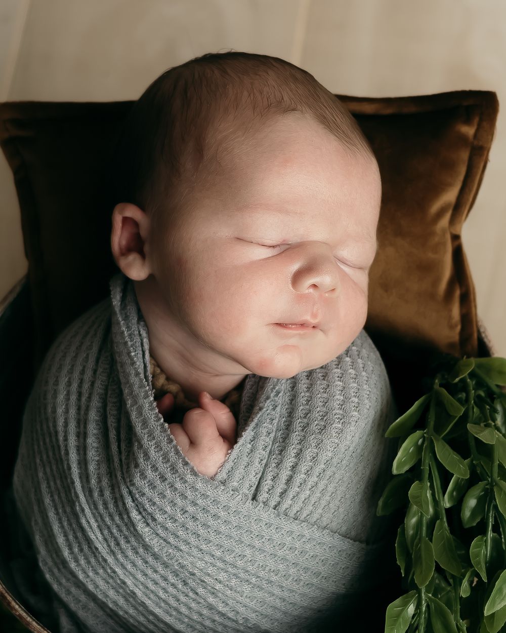 Sleeping newborn wrapped in grey knit blanket rests against brown pillow.