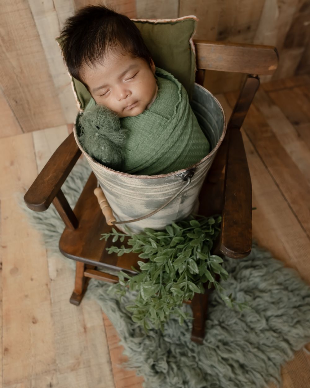 Sleeping baby wrapped in sage green blanket in vintage wooden chair with greenery.