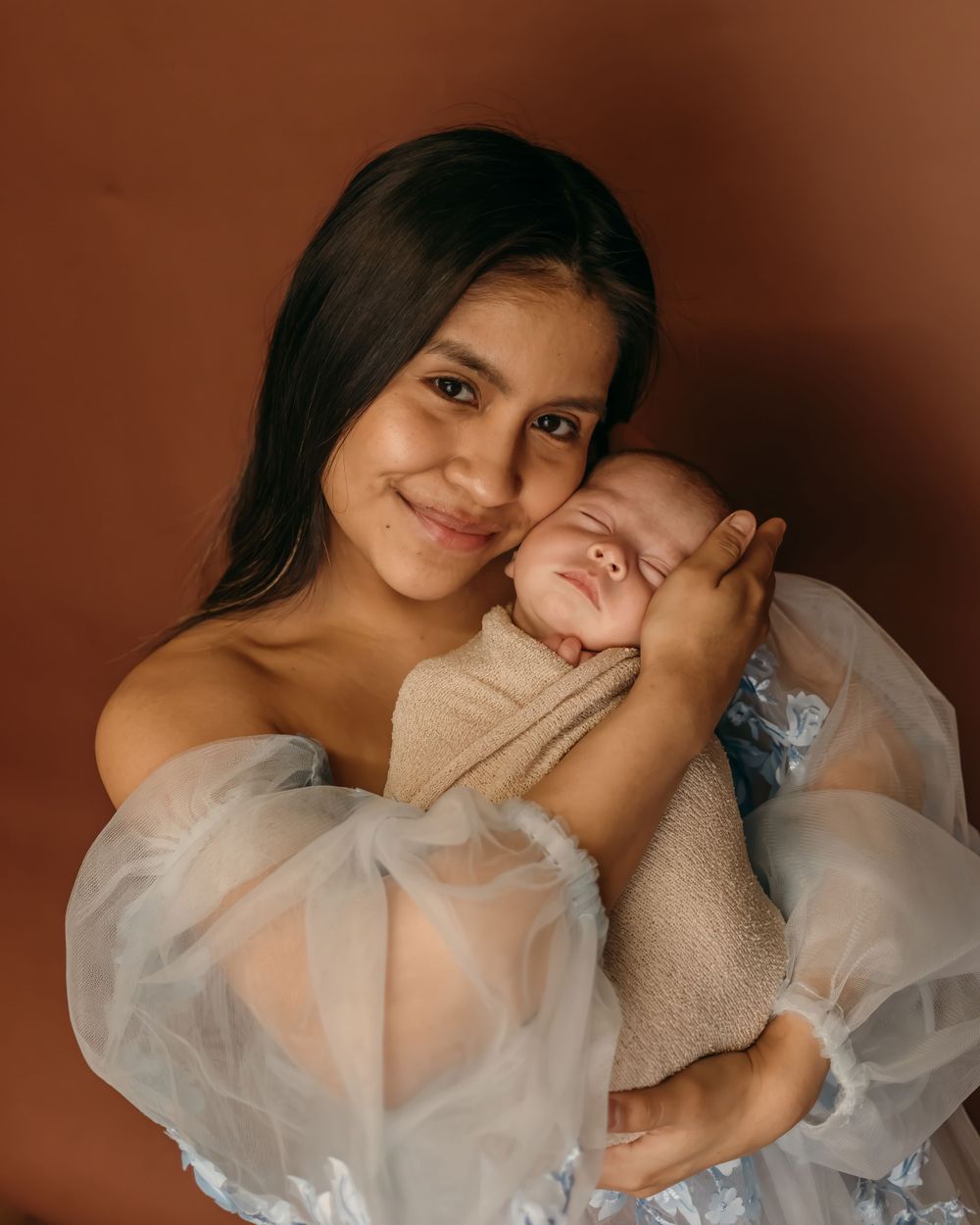 Tender moment between mother and sleeping baby in ethereal white dress.