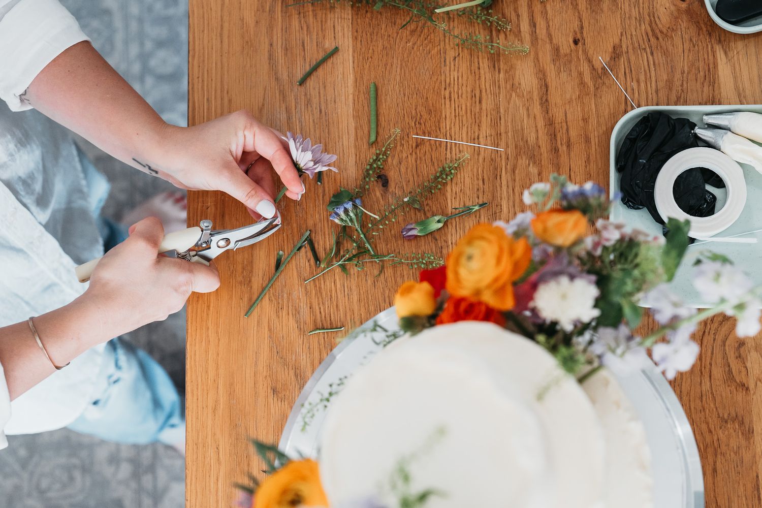 Hands trimming fresh flowers and greenery on wooden surface next to white plate with floral arrangement. Stow, Ohio