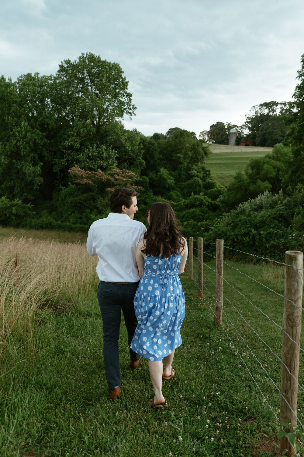 Couple walking together along a rustic fence line in a green countryside setting.