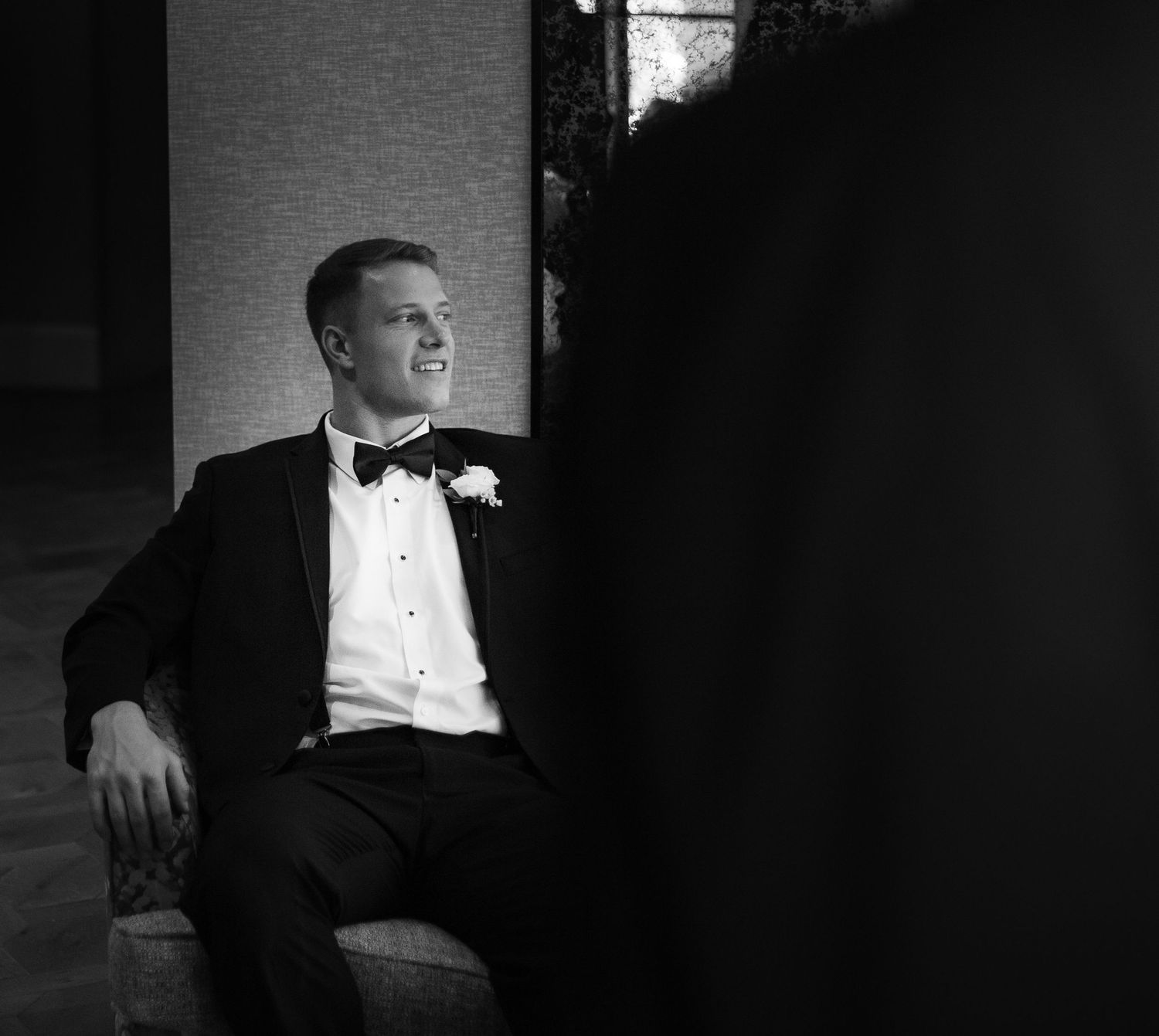Black and white photo of someone in a tuxedo and bow tie sitting by a window in dramatic lighting.