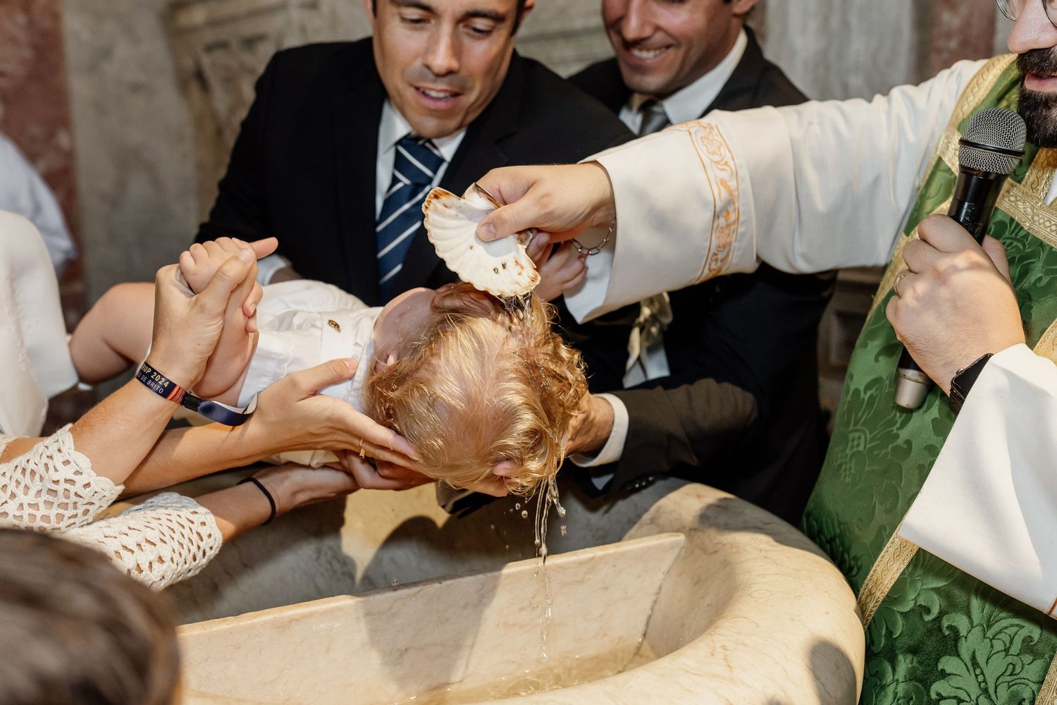 A priest performs a baptism ceremony by pouring holy water over a baby's head while family members gather around.