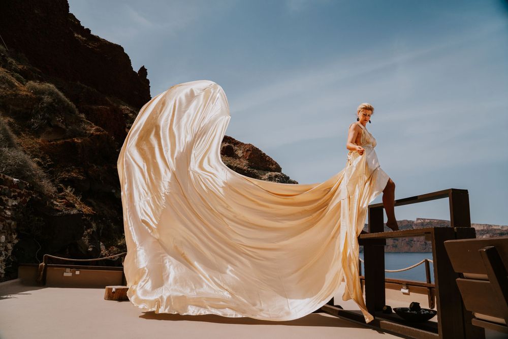 A flowing white wedding gown billows dramatically against coastal cliffs and Mediterranean waters.