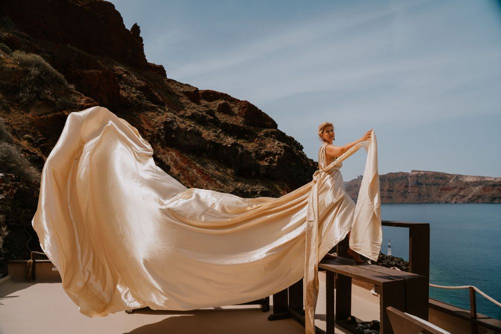 A white wedding dress catches the wind on a scenic Santorini terrace overlooking the sea.