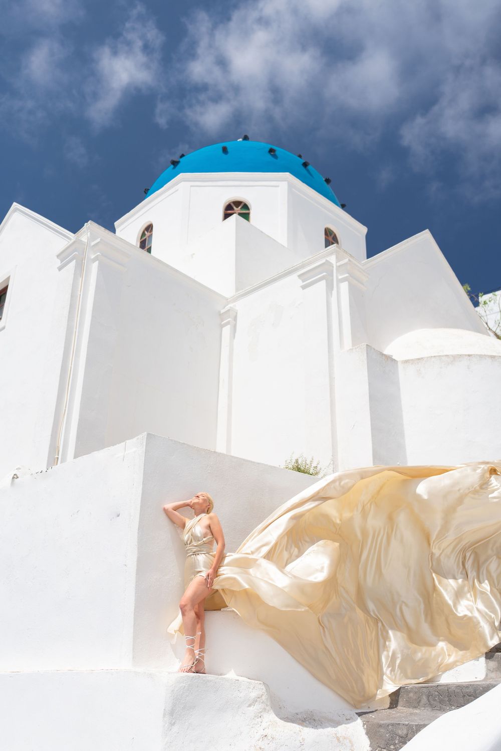 Ethereal golden dress flowing against traditional white Greek church architecture.