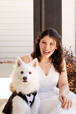 A bride in white dress poses with a fluffy white dog on a porch with pearl necklace.
