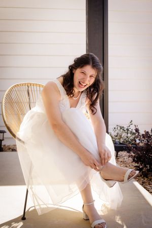 A bride in white adjusts her dress while sitting in a wicker chair on a porch.