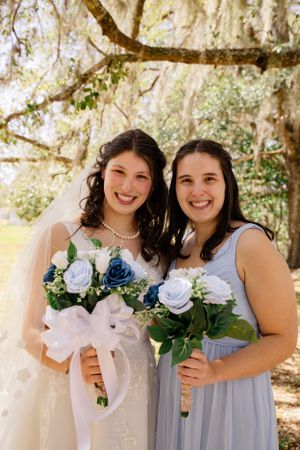 Wedding party members hold blue and white bouquets while posing under trees.