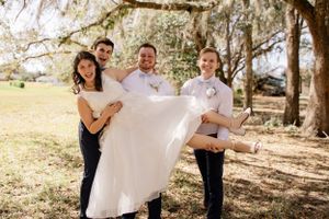 Wedding party members lift and carry bride in white dress under Spanish moss trees.