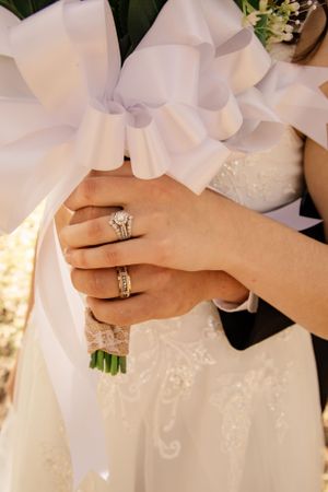 Close-up of hands wearing wedding rings with white dress and floral details.
