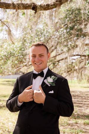 A groom in a black tuxedo smiles while reading a letter outdoors.