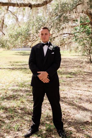 Groom in black tuxedo poses under Spanish moss draped trees in outdoor setting.