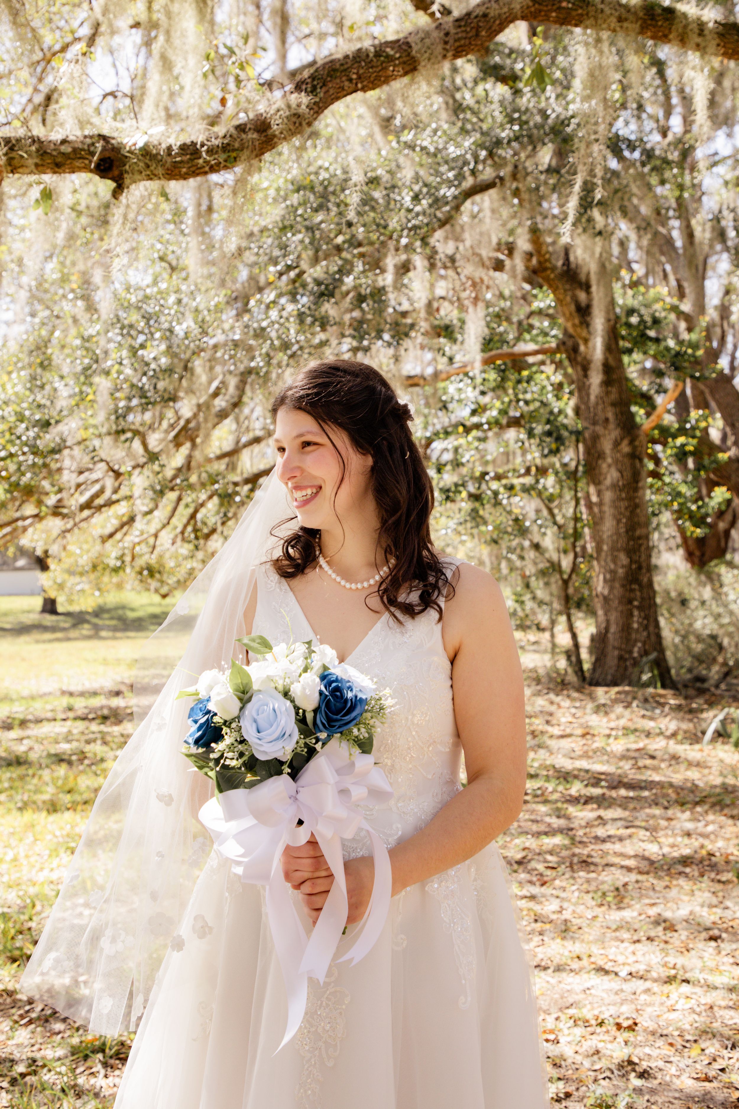 A bride in a white wedding dress poses under moss-draped trees holding a blue and white bouquet.