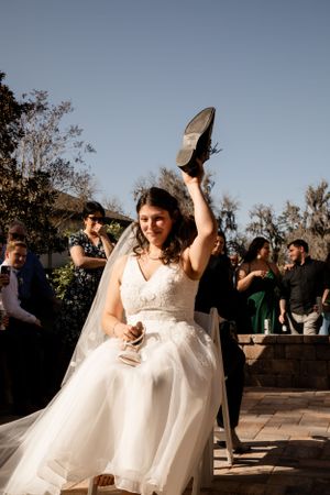 A bride in a white wedding dress holds up her shoe during a wedding game or tradition.