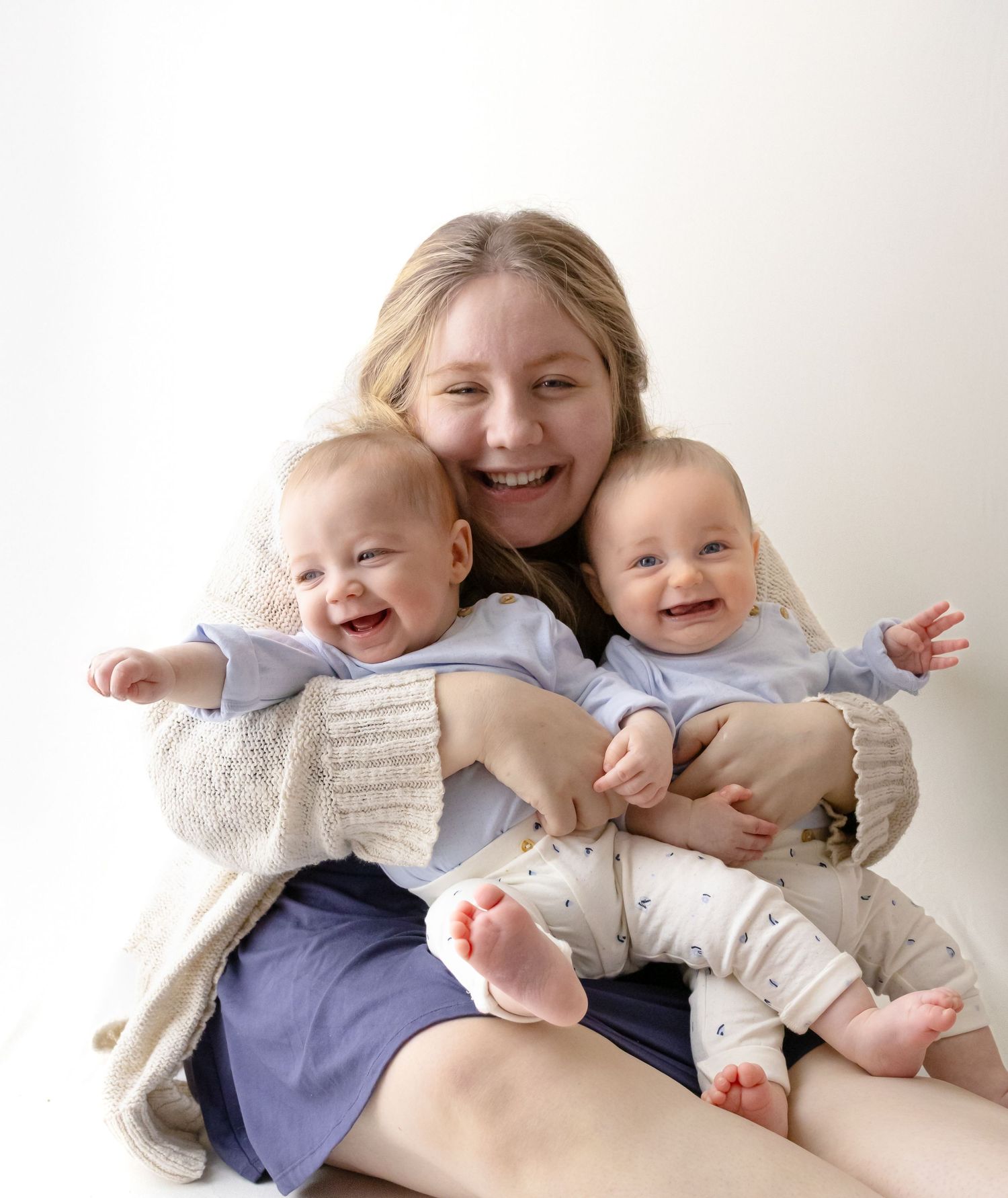 A mother and her twin boys sitting on the floor with a white backdrop, wearing blue tones. All are smiling and laughing