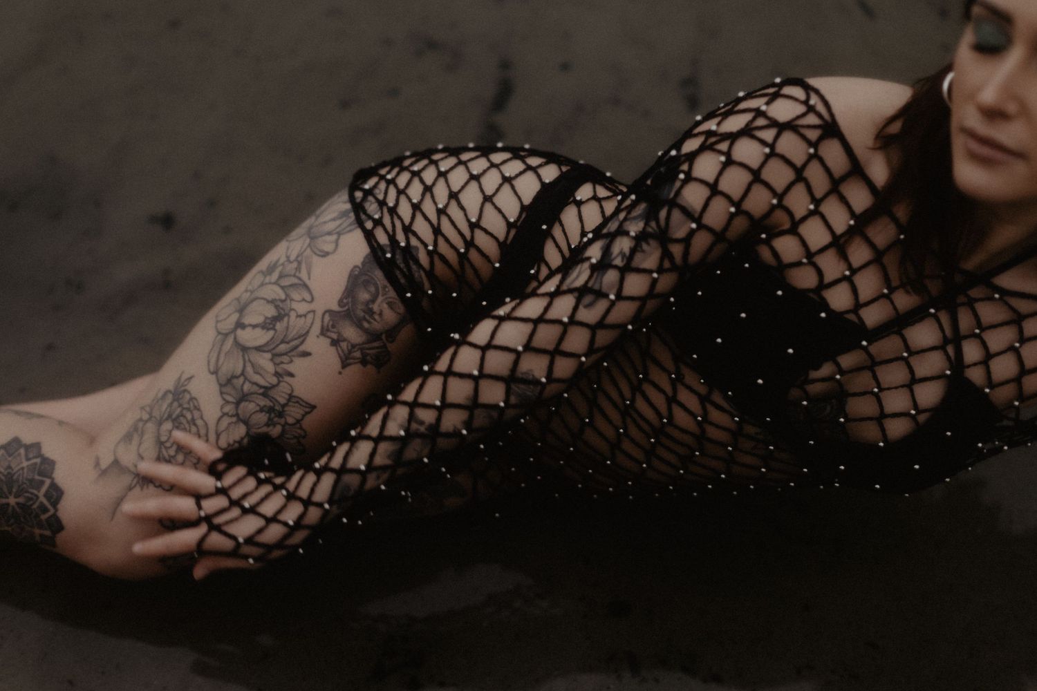 Figure in black fishnet bodysuit lounging artistically in moody dark setting.