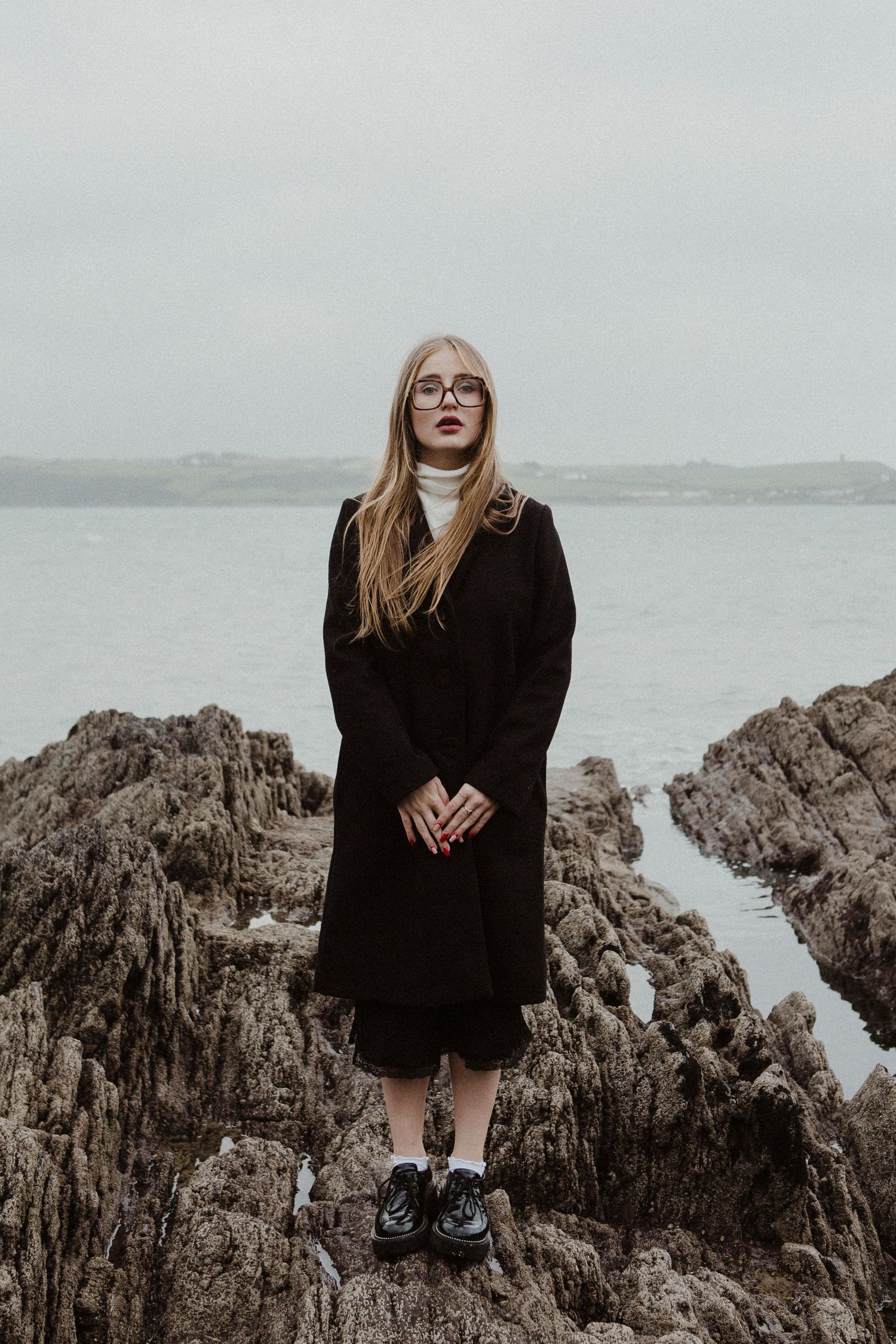 Person in black coat and turtleneck stands on coastal rocks with stormy ocean view behind.