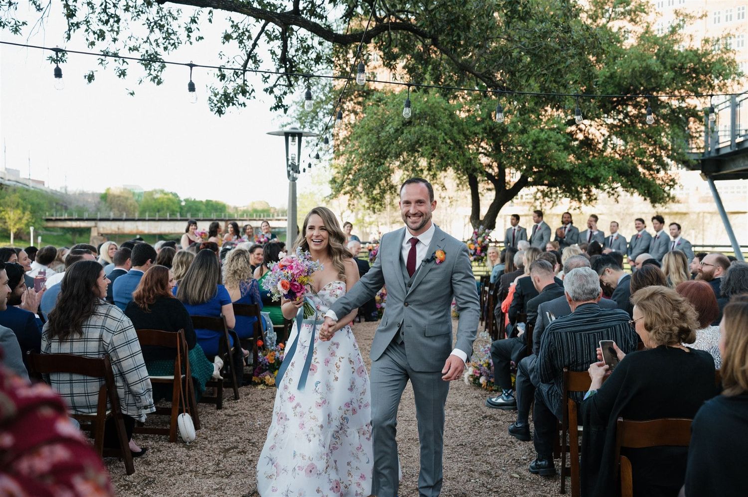 A newlywed couple walks down the outdoor aisle between seated guests during a garden wedding celebration.