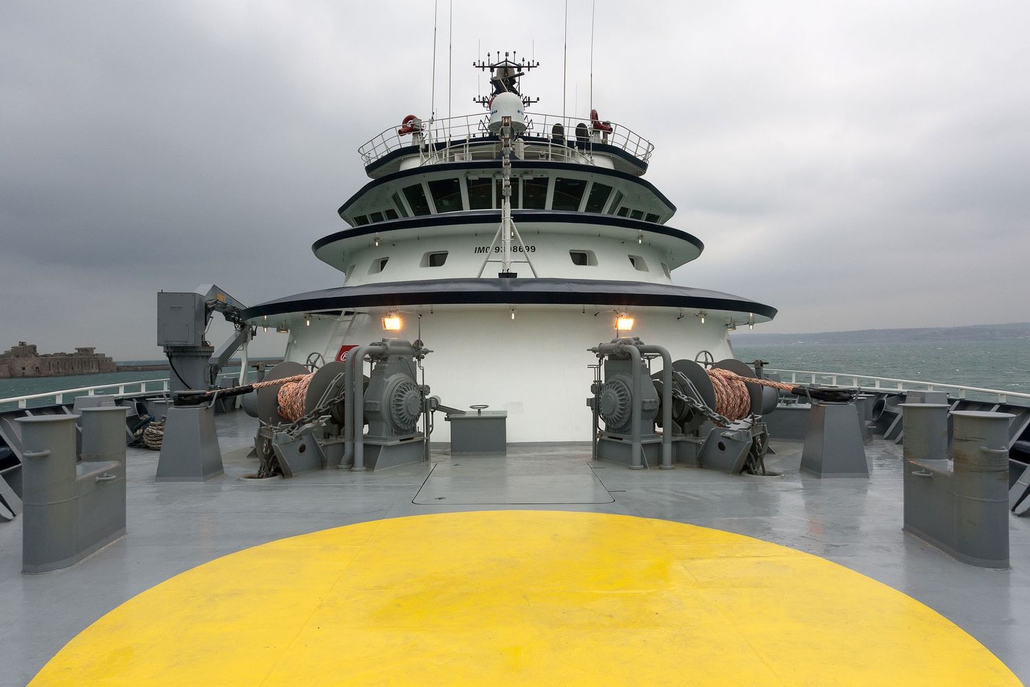 View from ship's bridge deck showing navigation equipment and yellow helipad under cloudy skies.