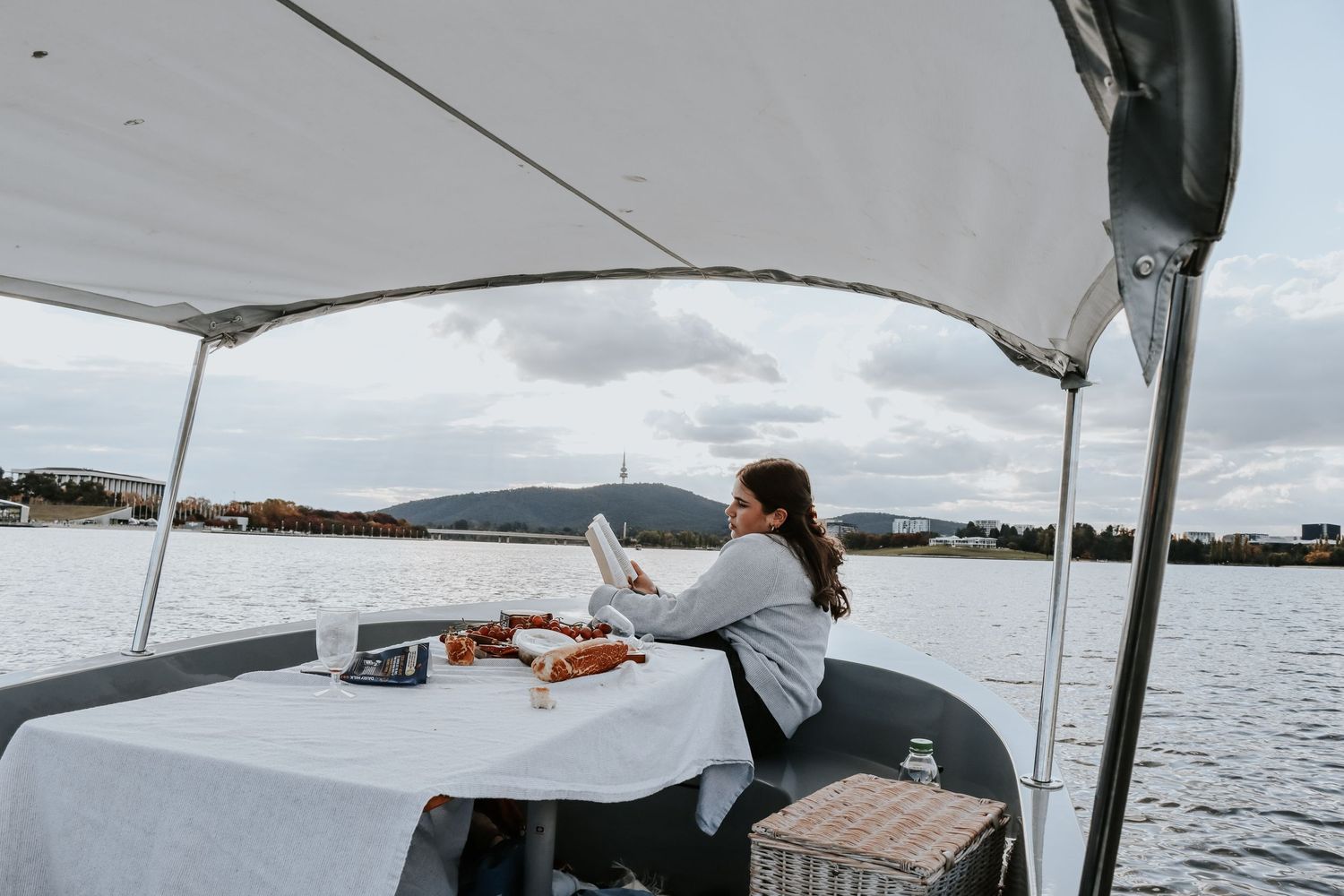Woman relaxing on a boat on a calm lake, reading while seated at a table with food and a picnic basket.