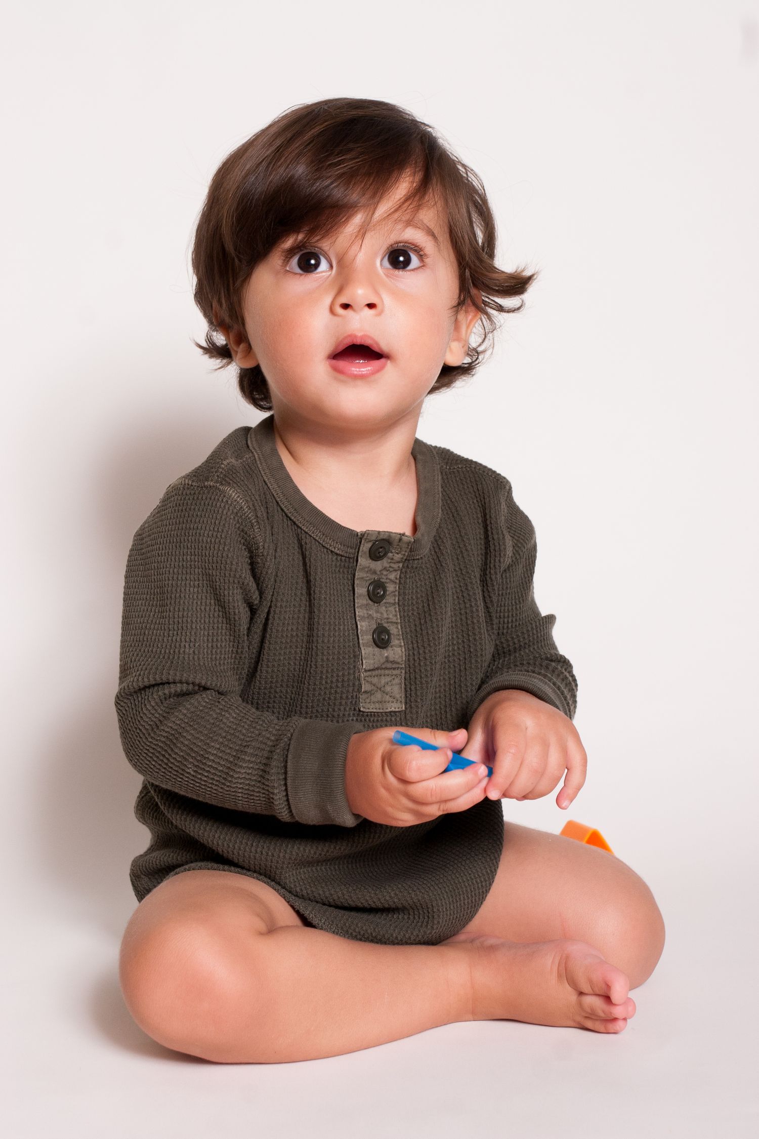 Toddler in grey henley shirt sitting on white background with surprised expression.