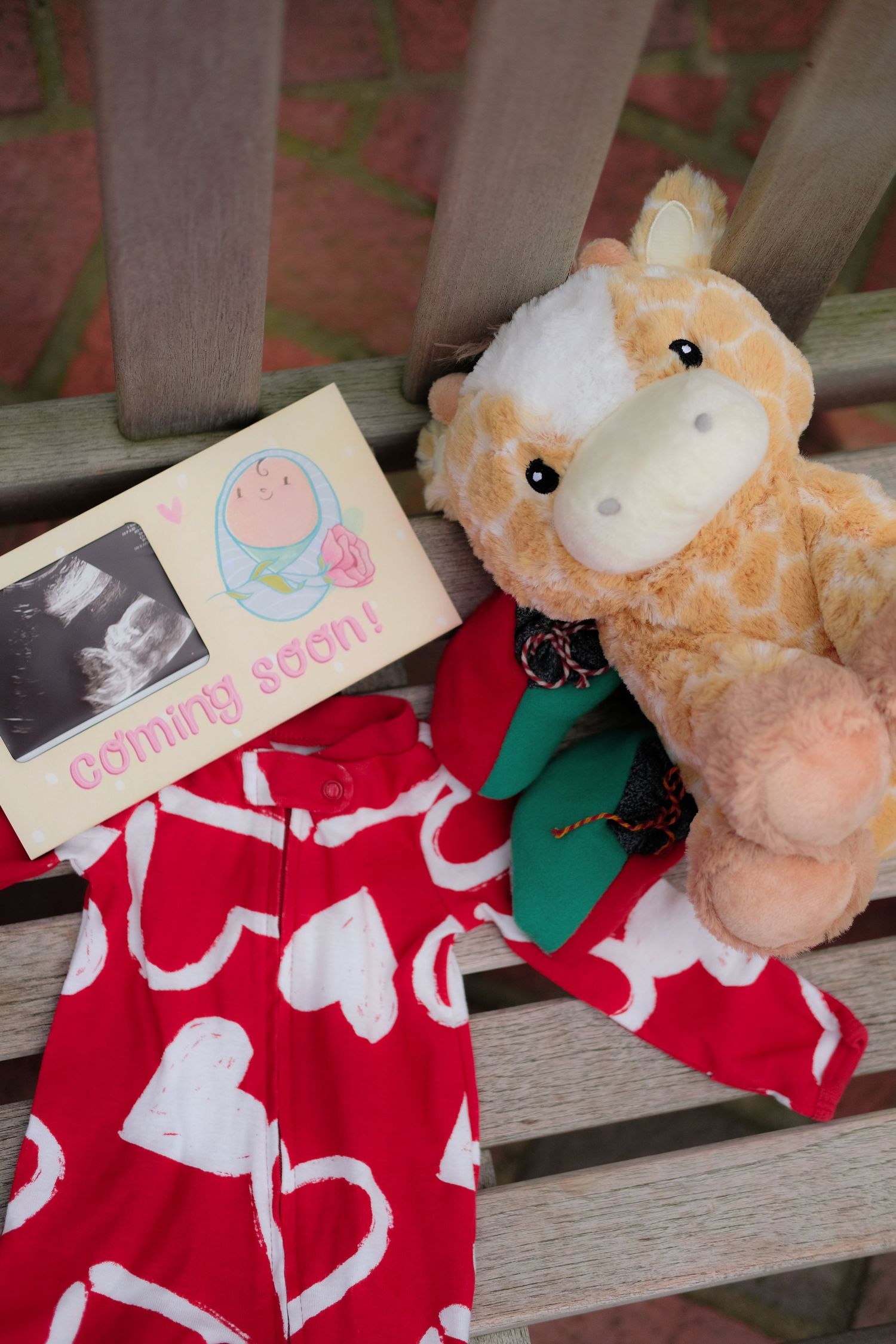 Baby announcement items including ultrasound photo, plush giraffe, and heart-patterned stocking on wooden bench.