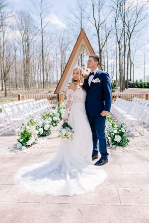 Wedding ceremony site features A-frame structure with white chairs and floral aisle decorations.