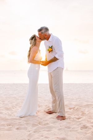 Romantic sunset beach wedding portrait of couple kissing in white attire.