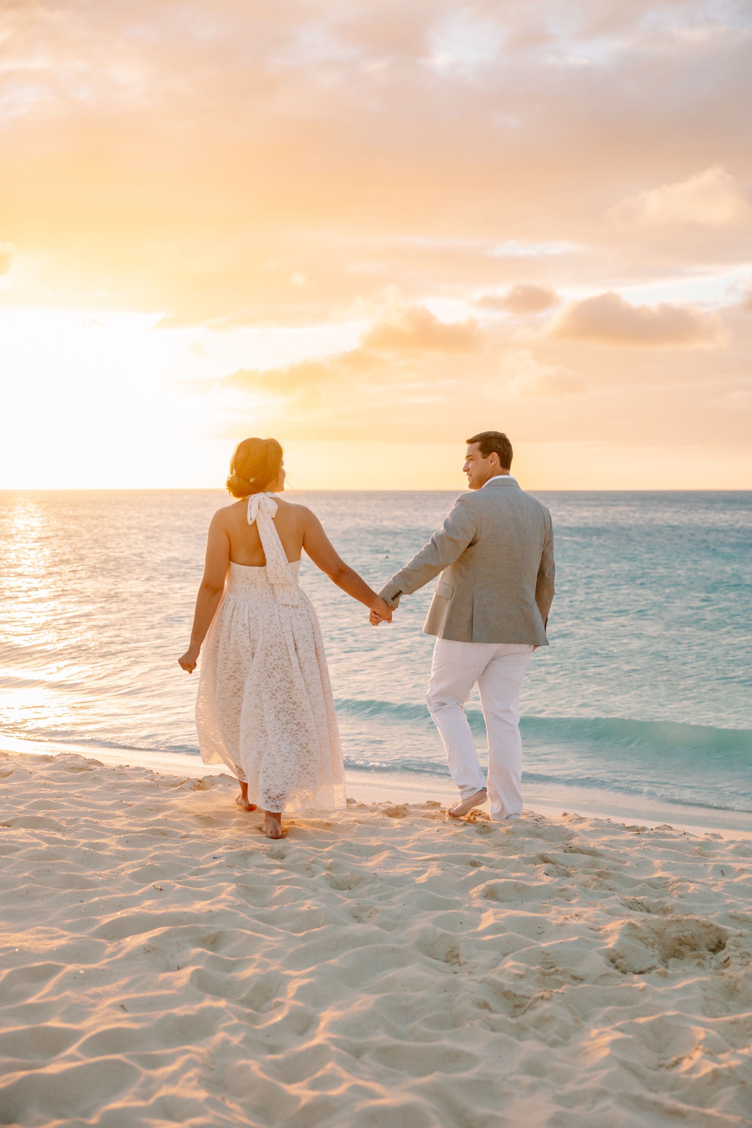 Couple in wedding attire holding hands walking on a sandy beach at sunset with turquoise ocean waves.