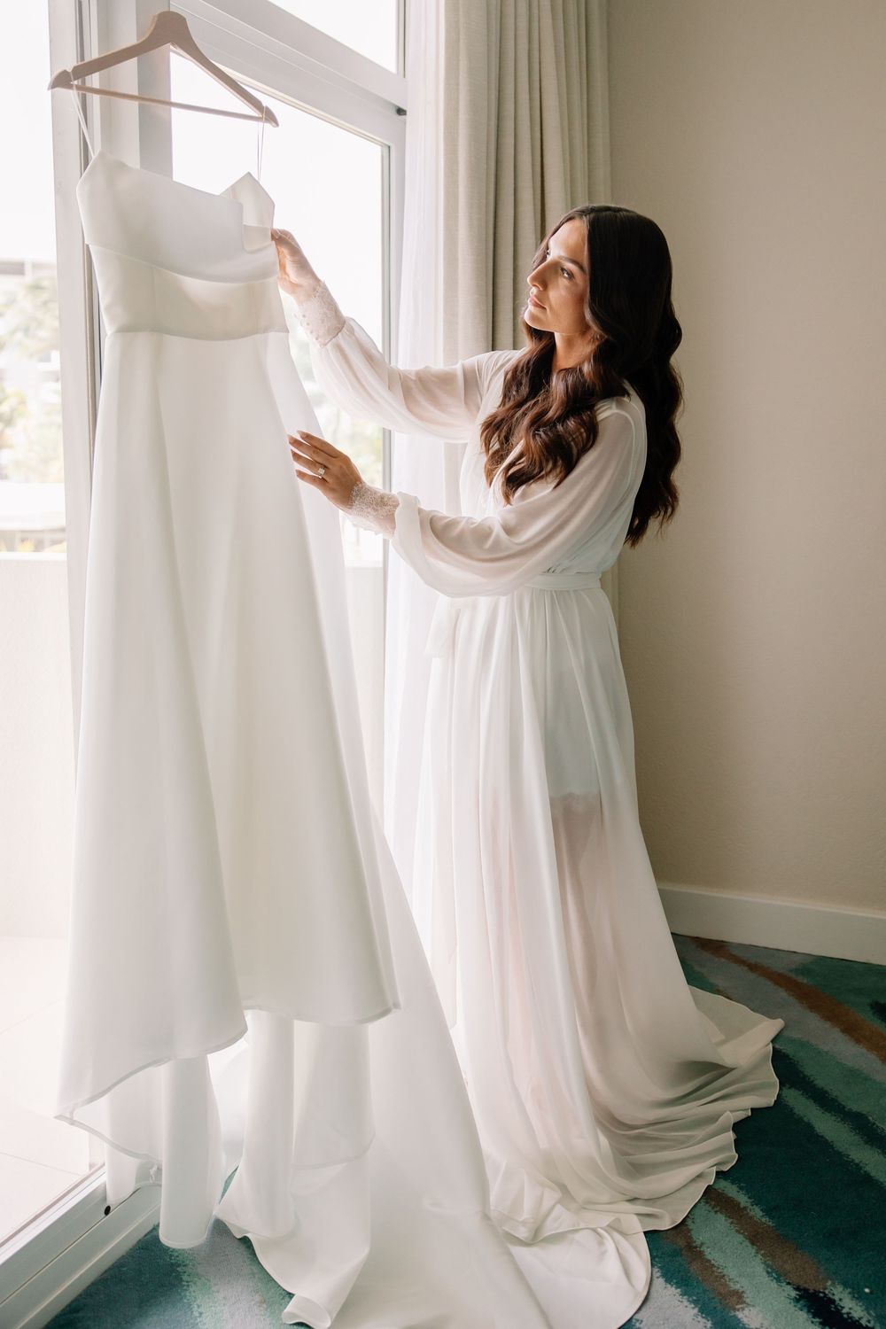 Wedding dress hanging by window as person in white robe admires it.