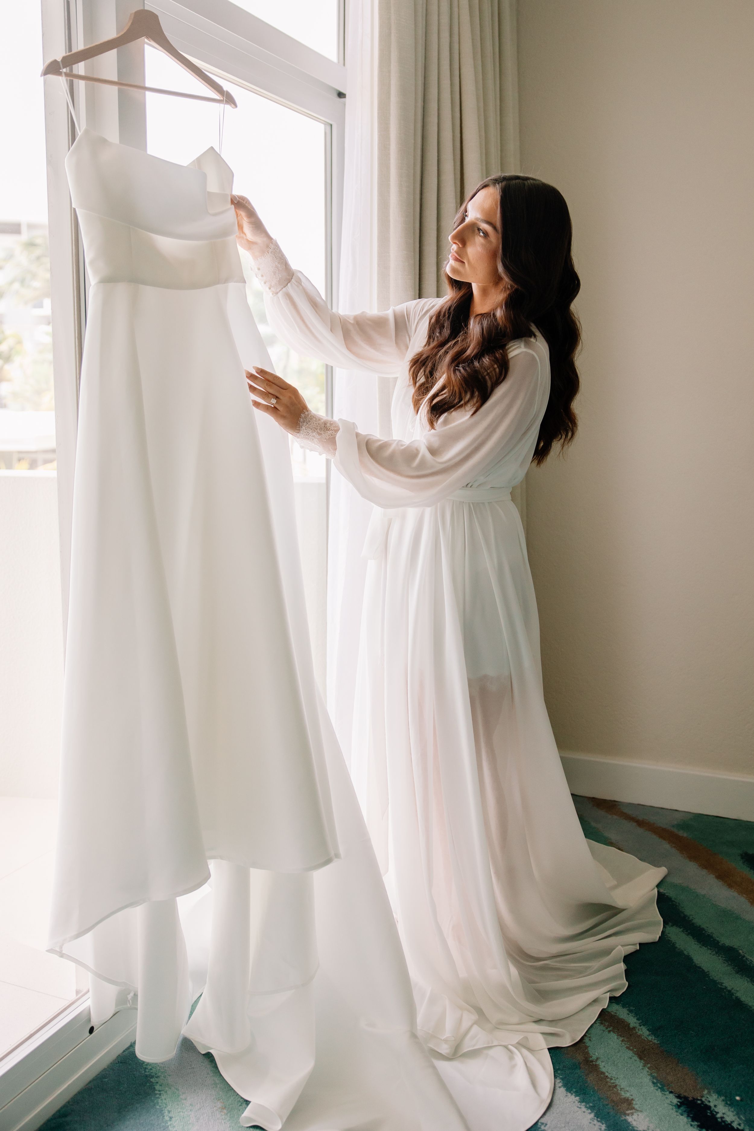 Wedding dress hanging by window as person in white robe admires it.