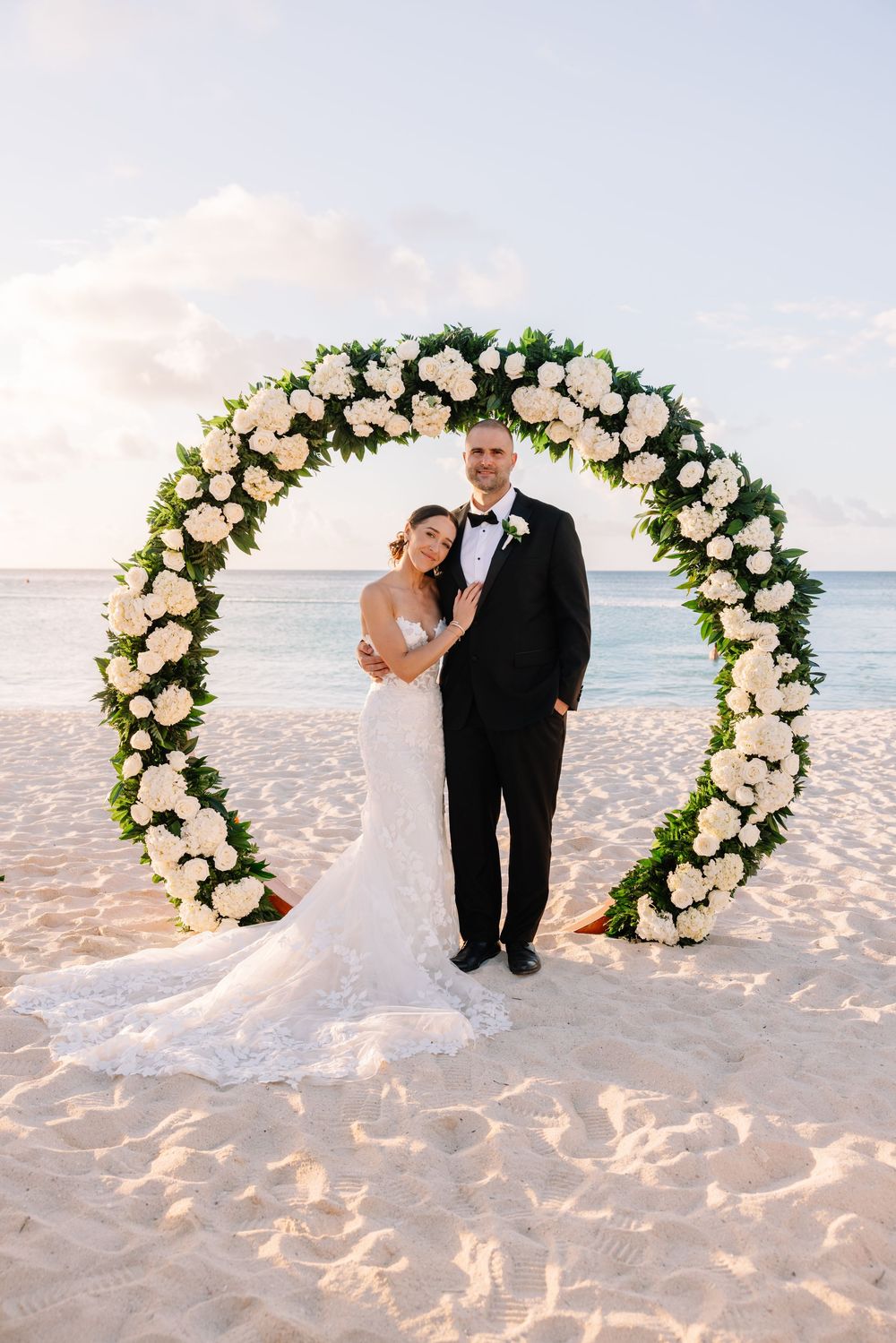 Bride and groom posing under a circular floral arch with white roses on a sandy beach at sunset.