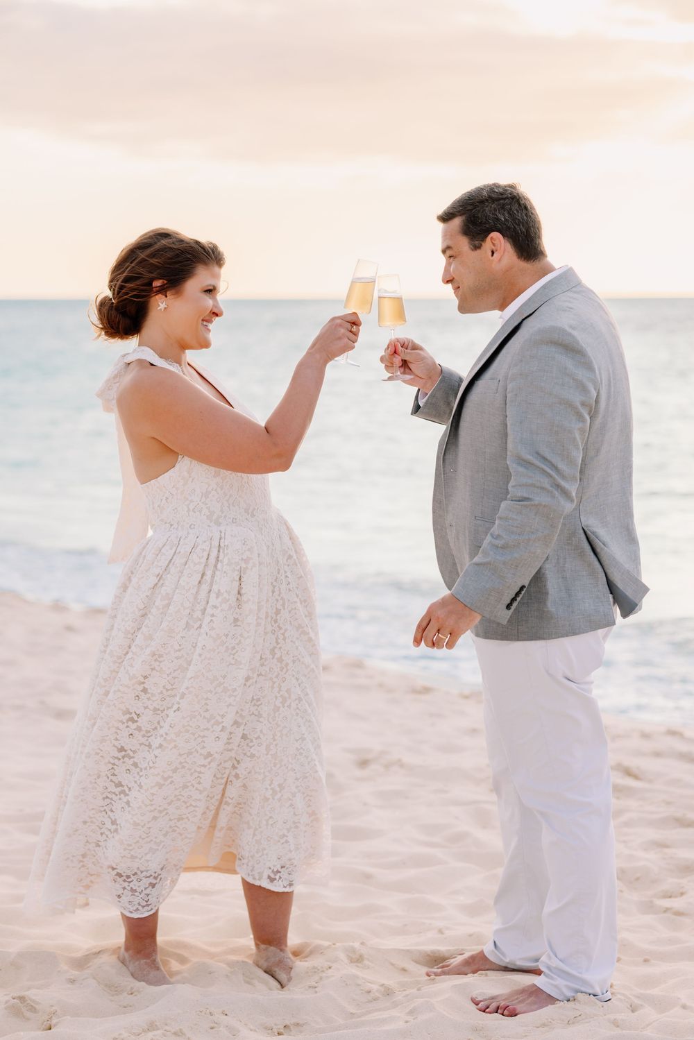 Couple toasting champagne glasses on a sandy beach at sunset, woman in white lace dress, man in gray suit.