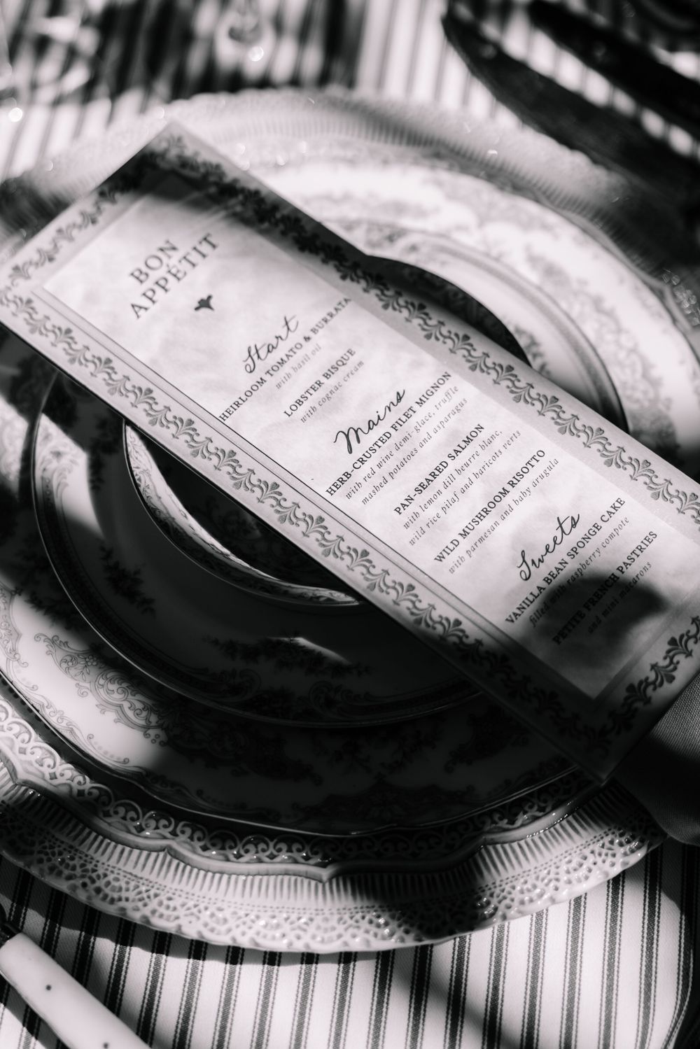 Black and white photo of an elegant place setting with a Bon Appetit menu card resting on stacked plates.