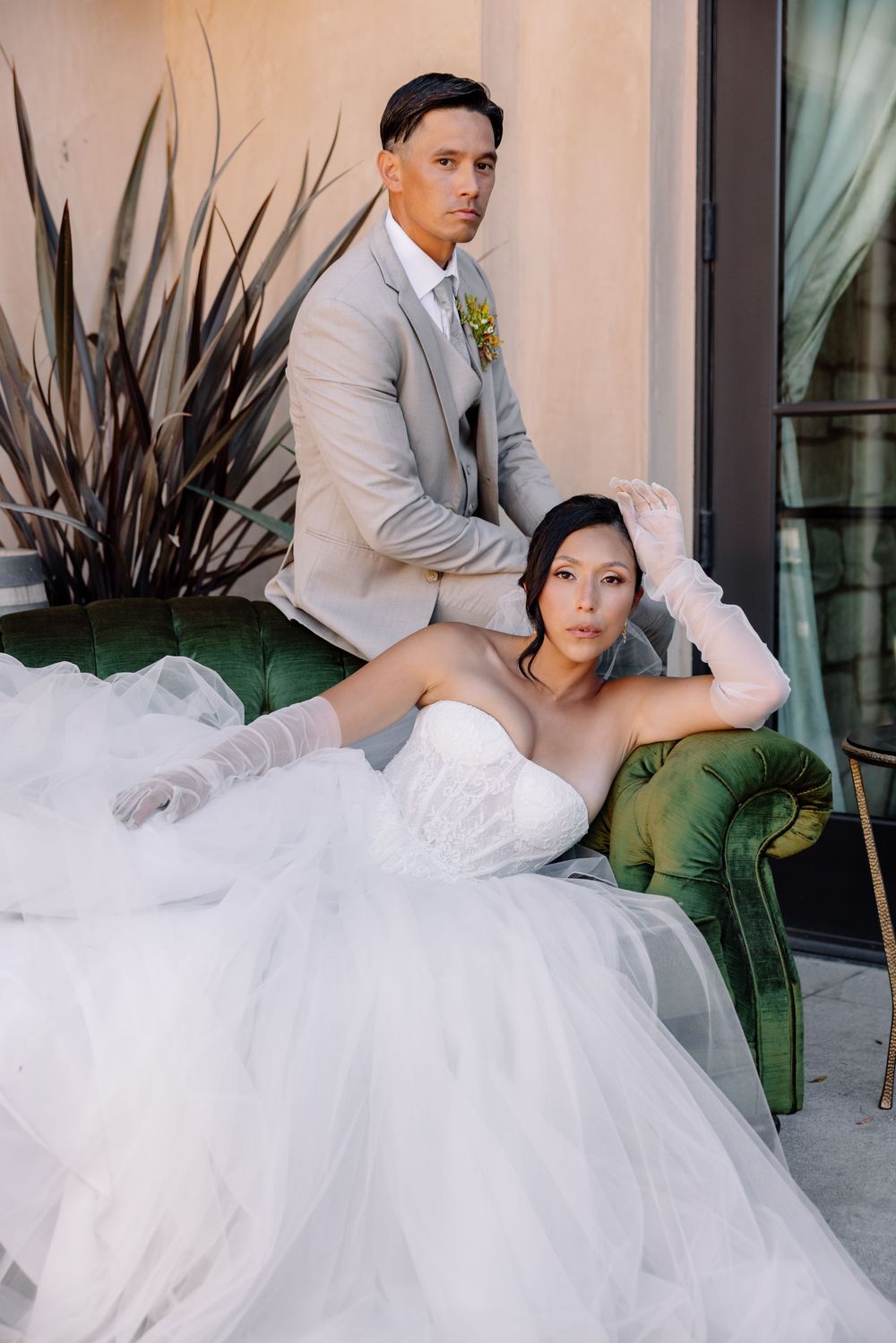 Bride in strapless white gown with gloves reclines on green velvet sofa as groom in gray suit stands behind her.