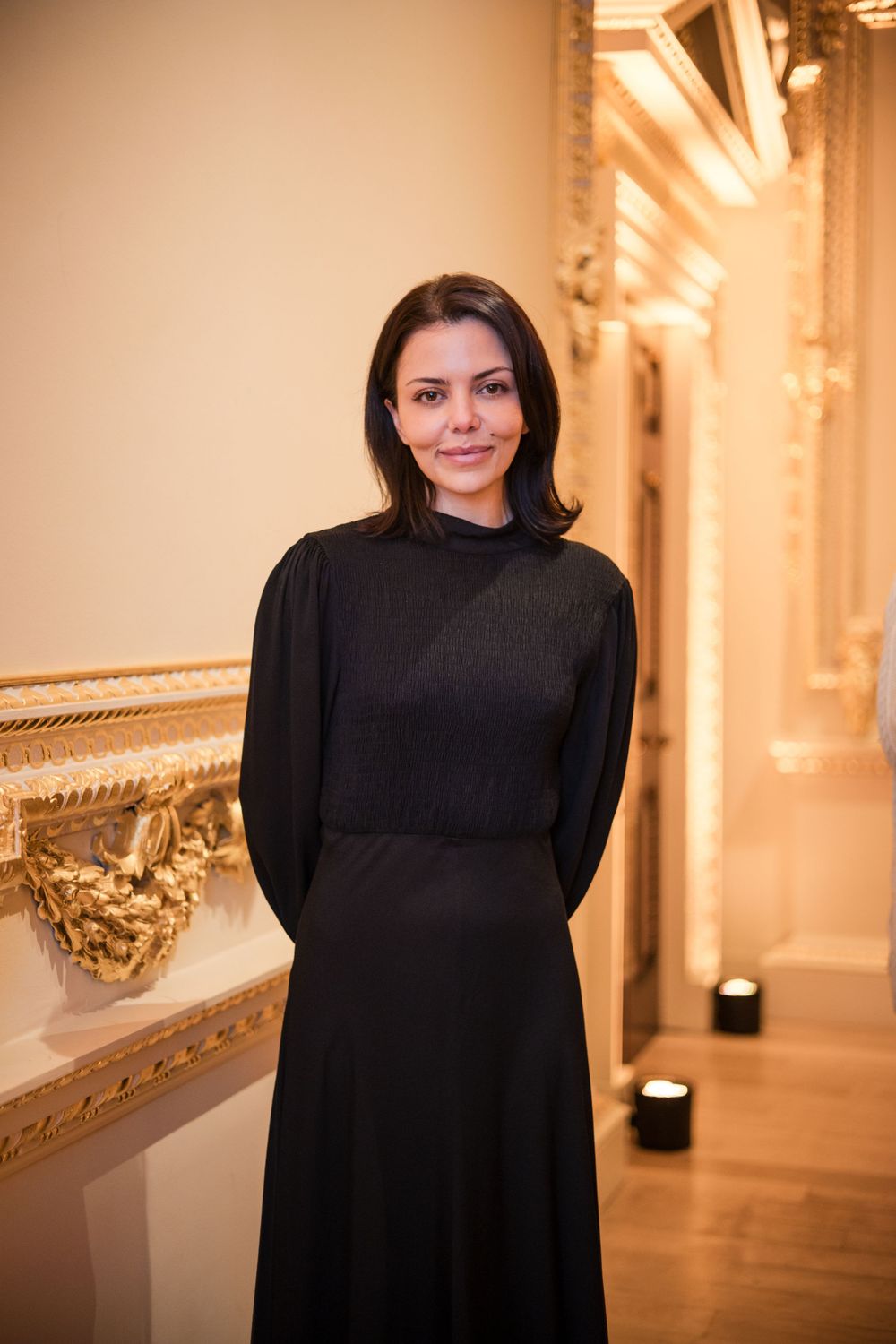 Elegant figure in black dress standing in ornate hallway with gold architectural details.
