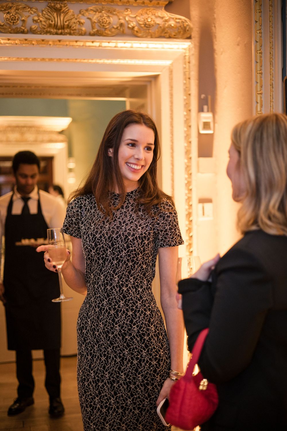 Elegant social gathering in ornate room with person in lace dress holding champagne.