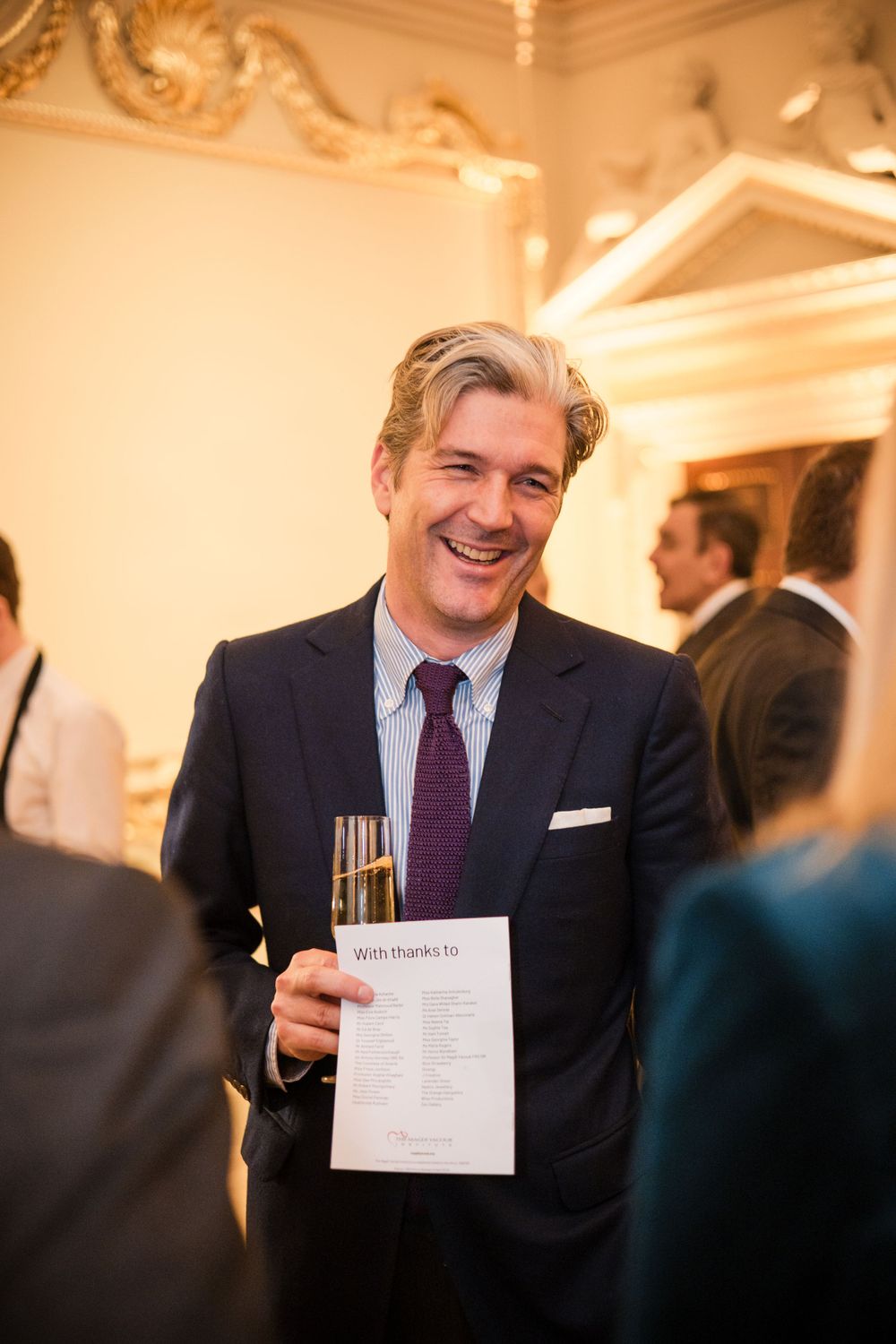 Formal event attendee in suit holding document and champagne in ornate venue.