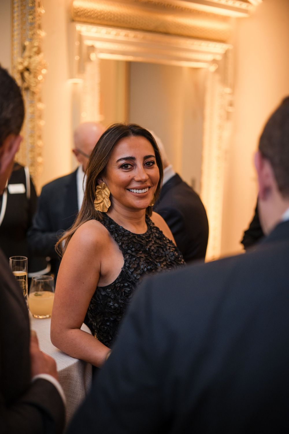 A woman in a black lace dress smiles warmly during an elegant indoor celebration.