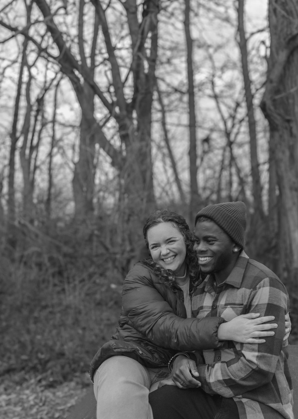 Black and white photo of a couple embracing and laughing in a wooded setting.